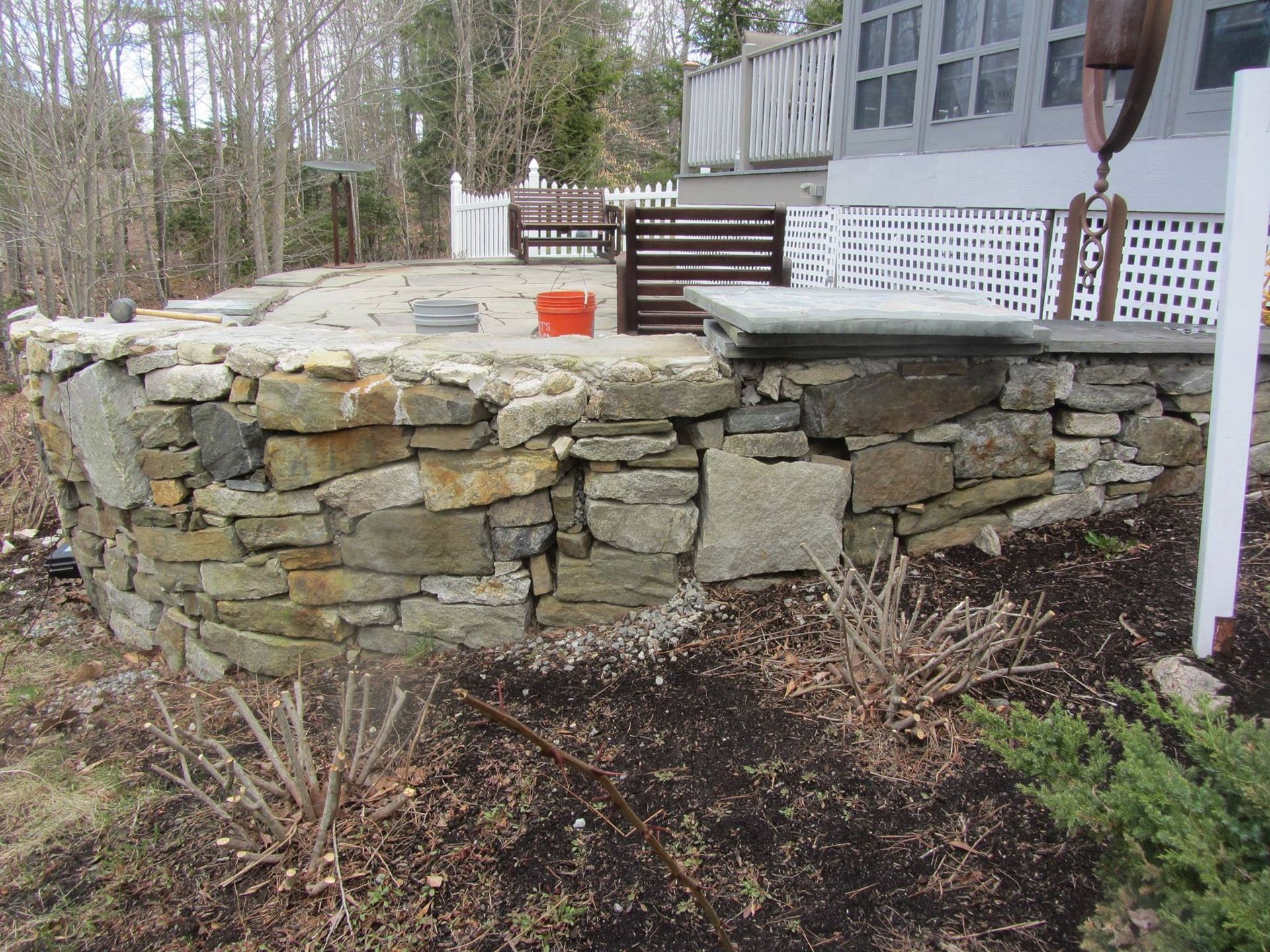 Stone retaining wall with a concrete patio beside a house and wooded yard