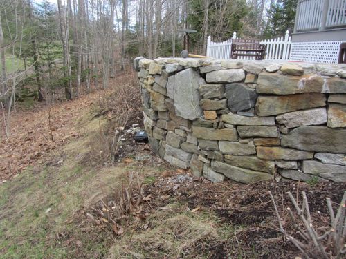 Stone retaining wall beside a house on a wooded hillside with bare trees and brown leaves