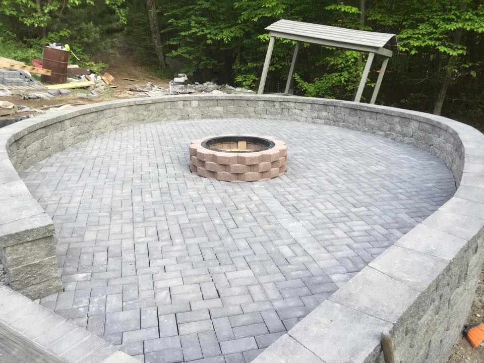Circular brick fire pit area with a central stone ring, surrounded by a curved retaining wall outdoors.