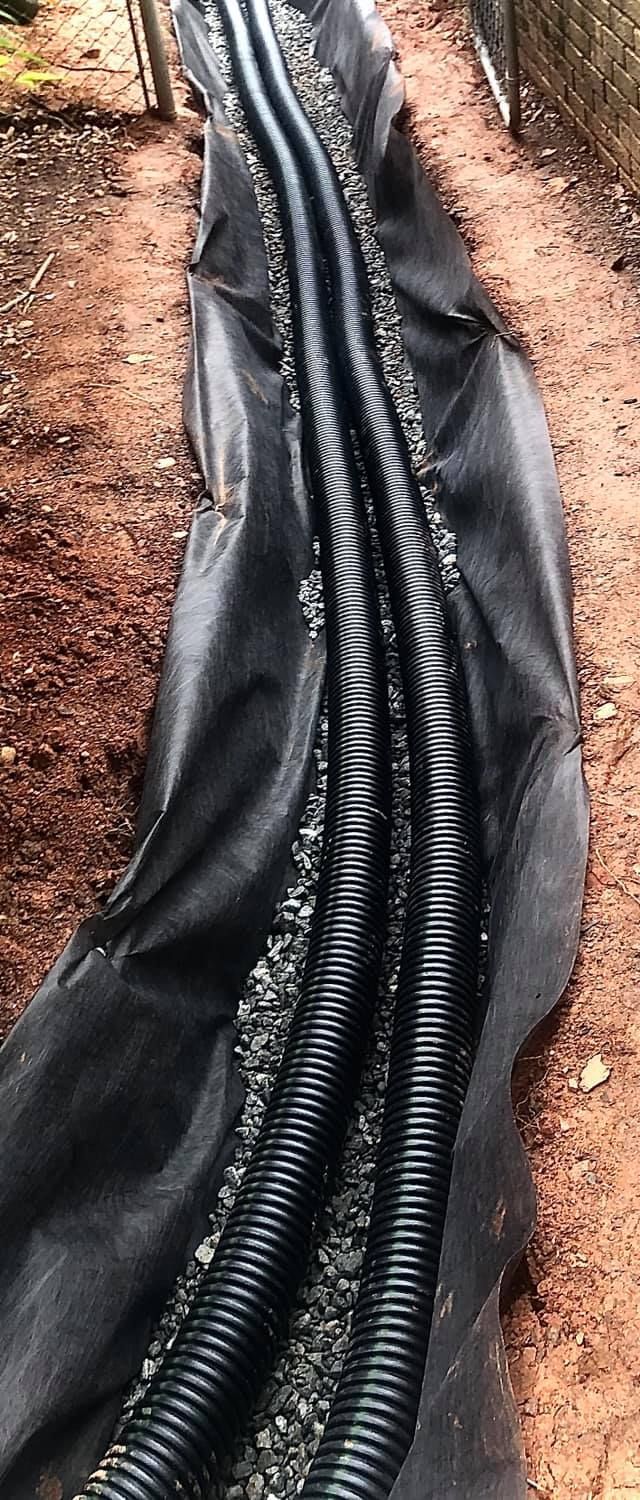 A trench lined with black landscape fabric, containing gravel and two parallel lengths of black corrugated drainage pipe.