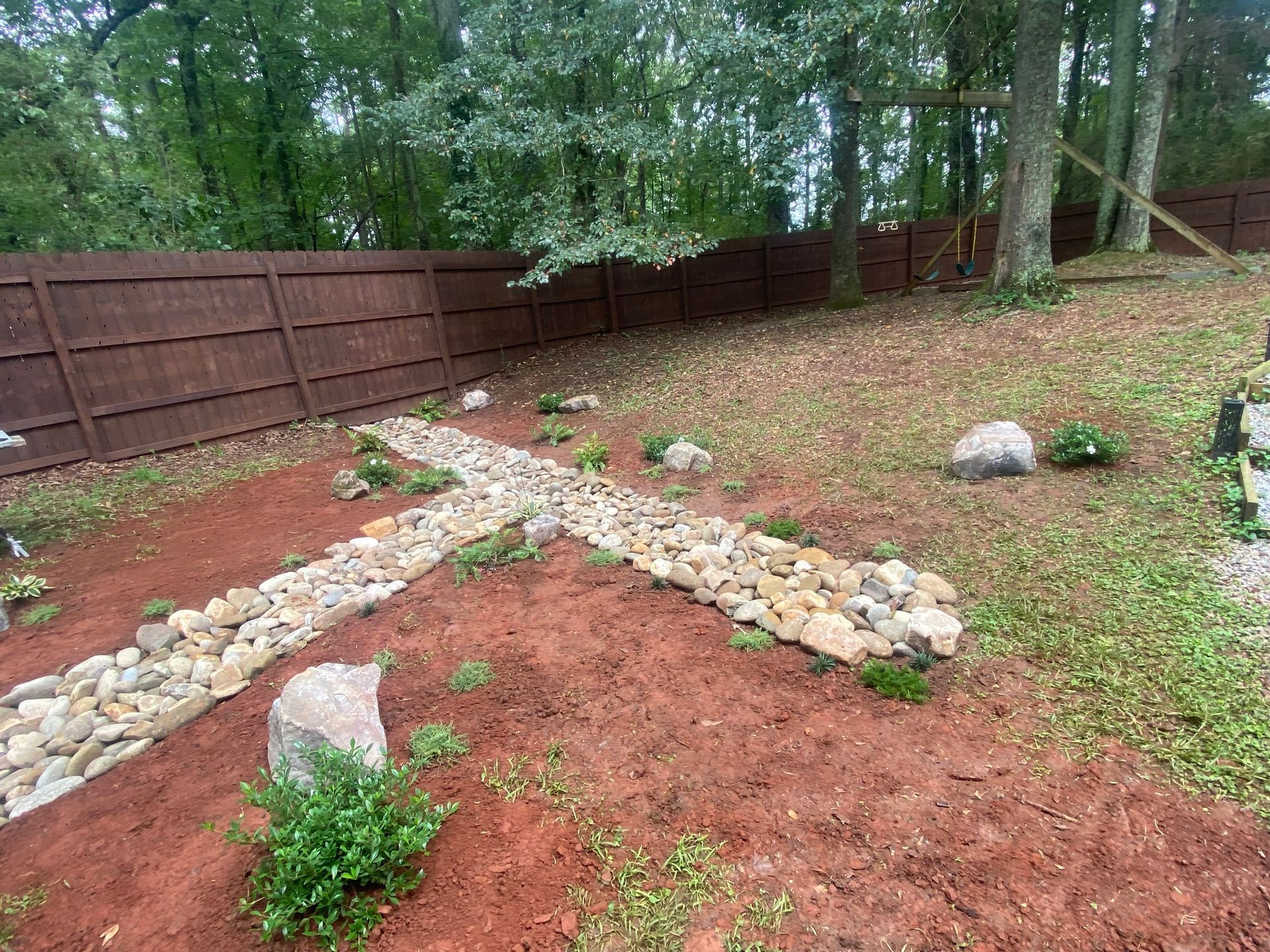 A stone-lined path shaped like a cross in a backyard with red mulch, scattered plants, and a brown wooden fence.
