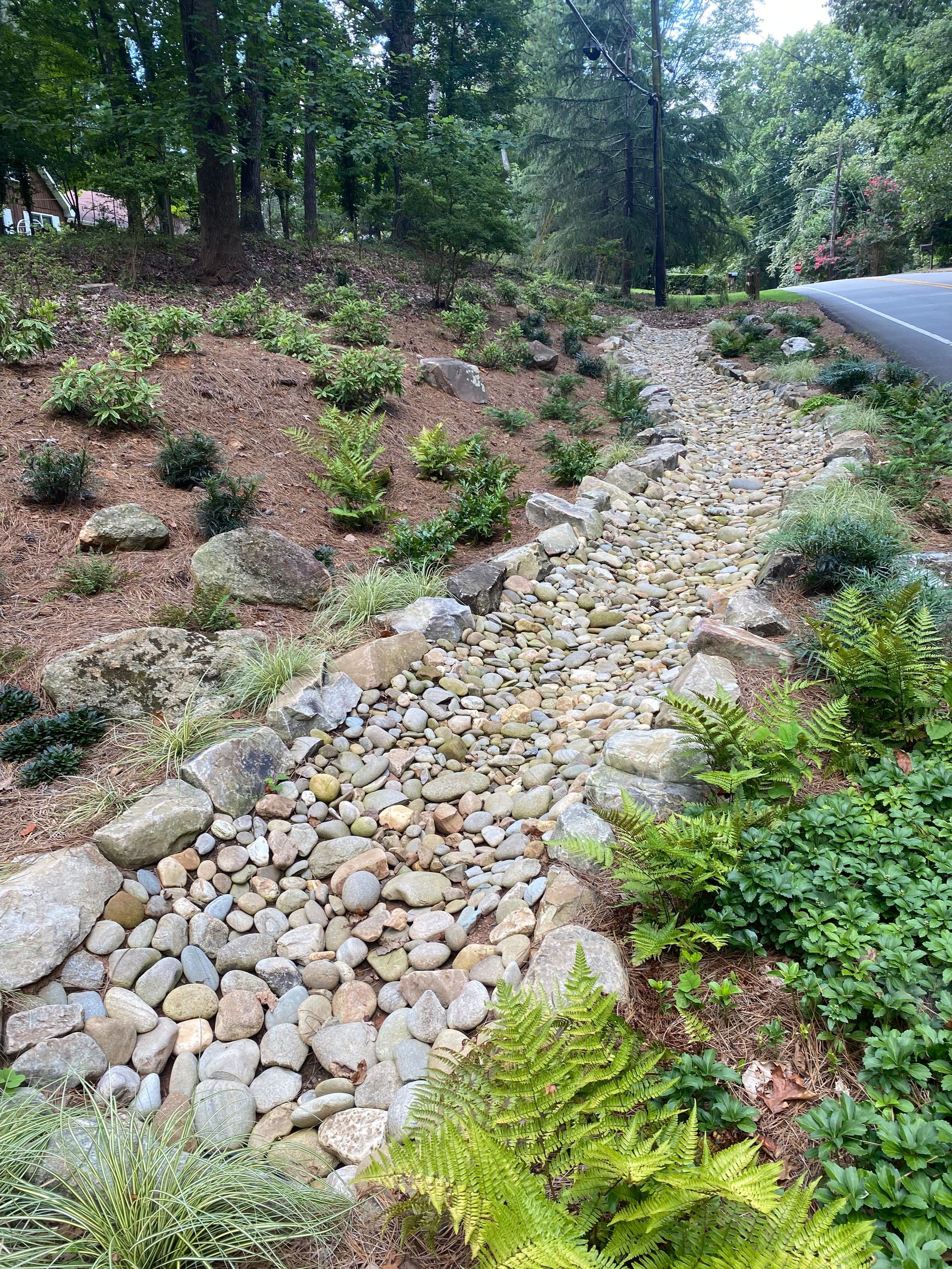 A drainage grate runs alongside a strip of red gravel, bordering green grass on one side and a stone walkway on the other. A drainage grate runs alongside a strip of red gravel, bordering green grass on one side and a stone walkway on the other.