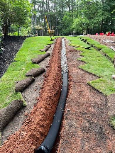 Black drainage pipe in a red dirt trench between green grass strips at a park or construction site.