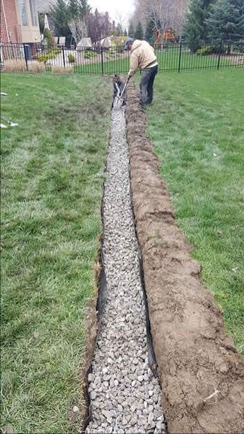 Narrow gravel trench runs through a grassy yard beside a dirt mound and fence.