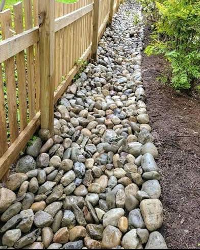Rock-filled drainage trench beside a wooden fence, with soil and shrubs along the edge.