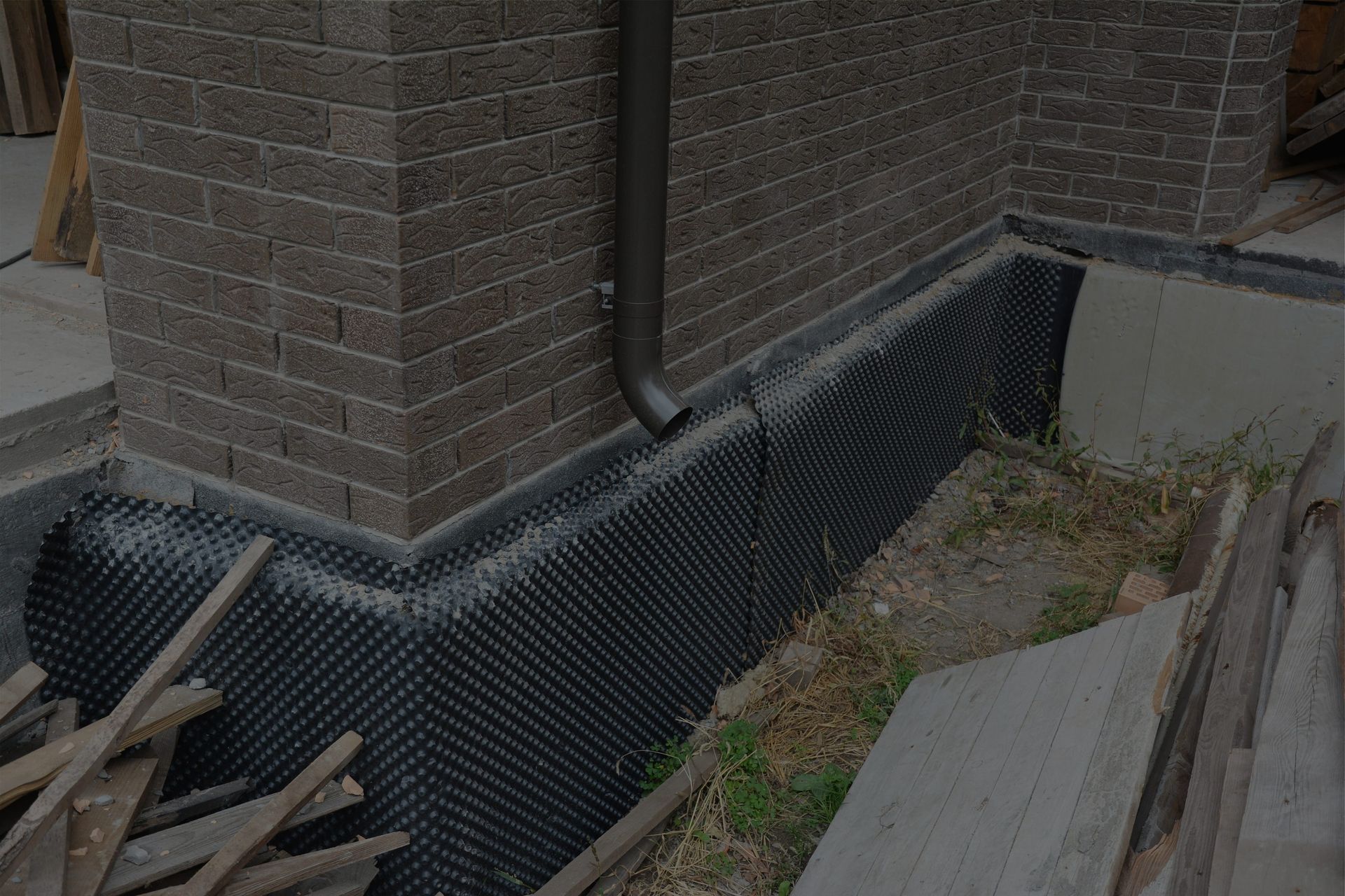 Black textured foundation wrap on brick house corner beside concrete slab and scattered boards