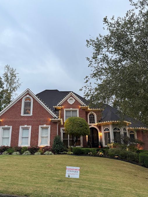 A two-story red brick house with a dark roof and white trim, decorated with string lights, sitting on a grassy lawn. A two-story red brick house with a dark roof and white trim, decorated with string lights, sitting on a grassy lawn.