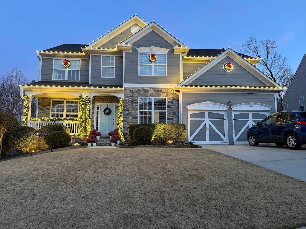 A two-story gray house with stone accents, decorated with Christmas lights and wreaths, under a twilight sky.