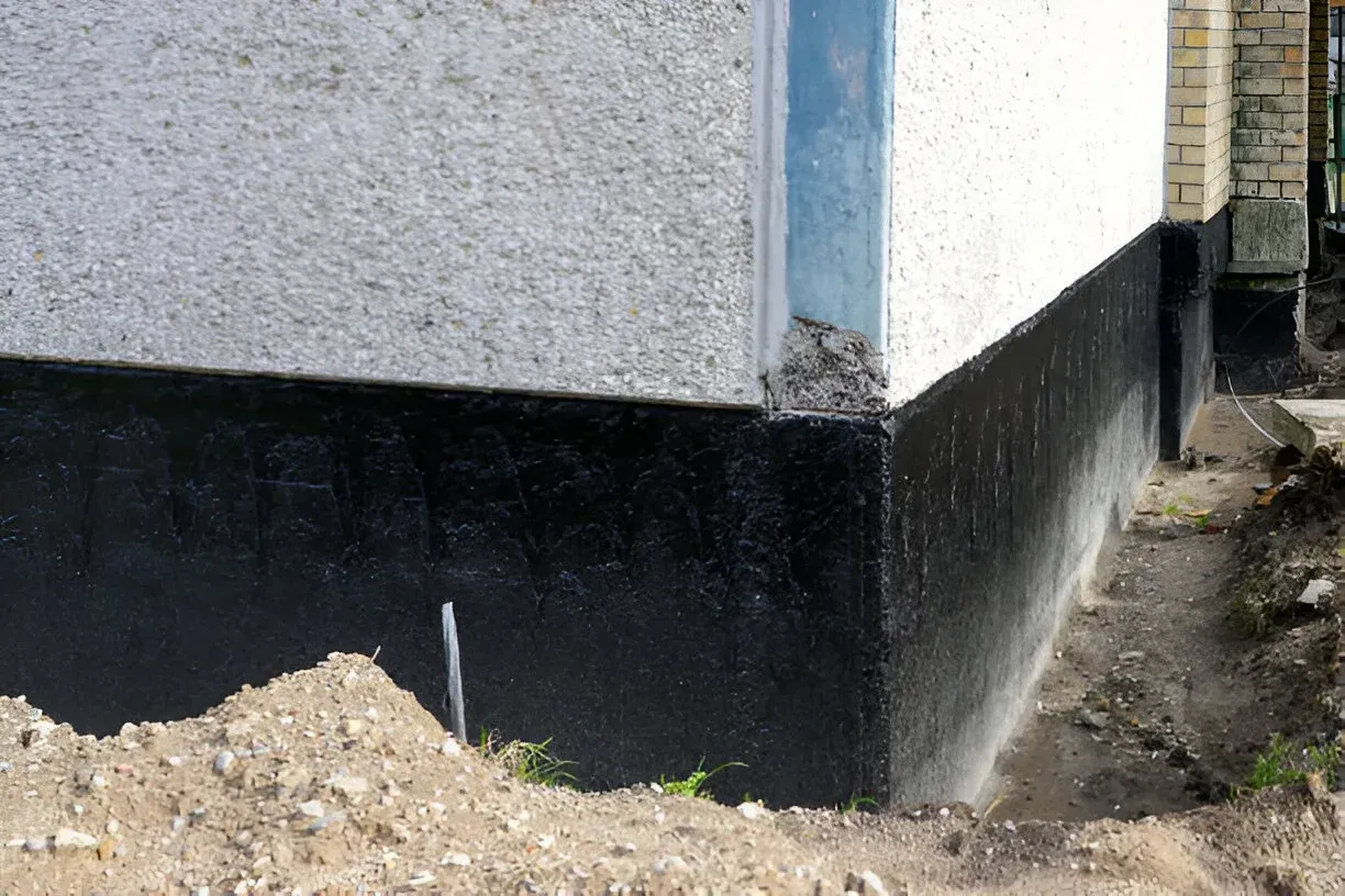 A building corner with a white stucco wall above a black, tar-coated foundation base, next to a trench of excavated soil.