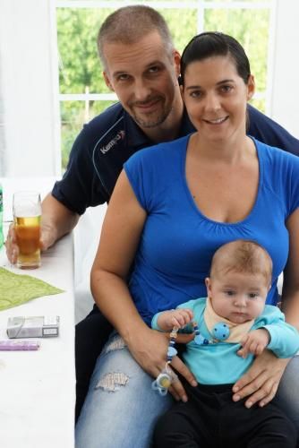 Eine Frau in einem blauen Hemd hält ein Baby, während ein Mann ein Glas Bier hält