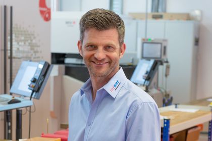 Ein Mann in einem blauen Hemd lächelt vor einer Maschine.