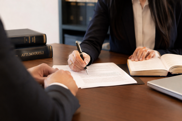 Due persone, sedute a una scrivania, discutono di un documento, utilizzando penna, taccuino e computer portatile, durante una riunione di lavoro.