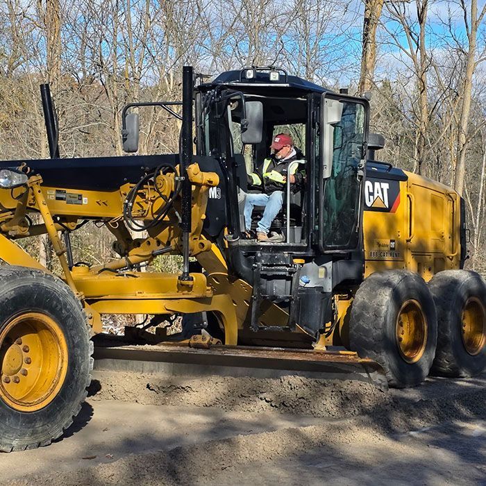 Yellow CAT construction vehicle with operator in cab on a dirt road near bare trees