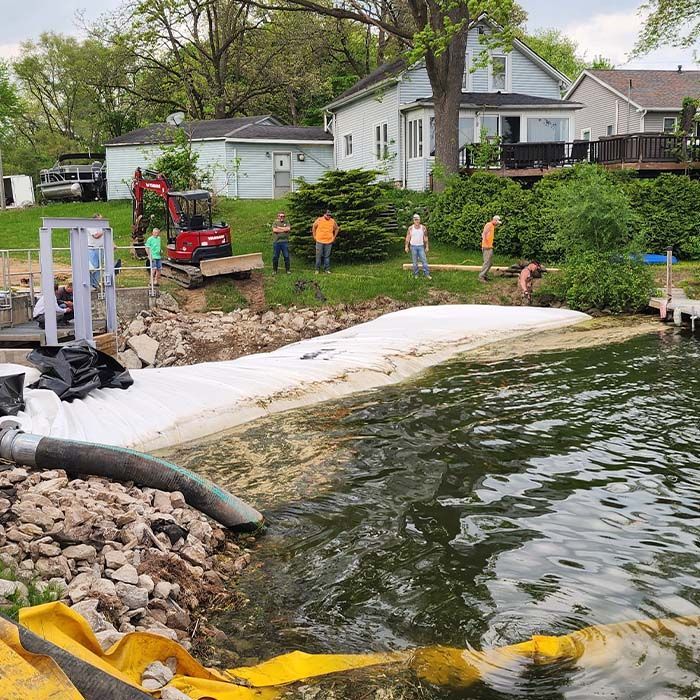 Shoreline repair with workers, rocks, and white erosion blanket beside a lakeside house