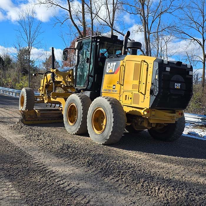 Yellow CAT motor grader on a gravel road under a blue sky, with bare trees and patches of snow.