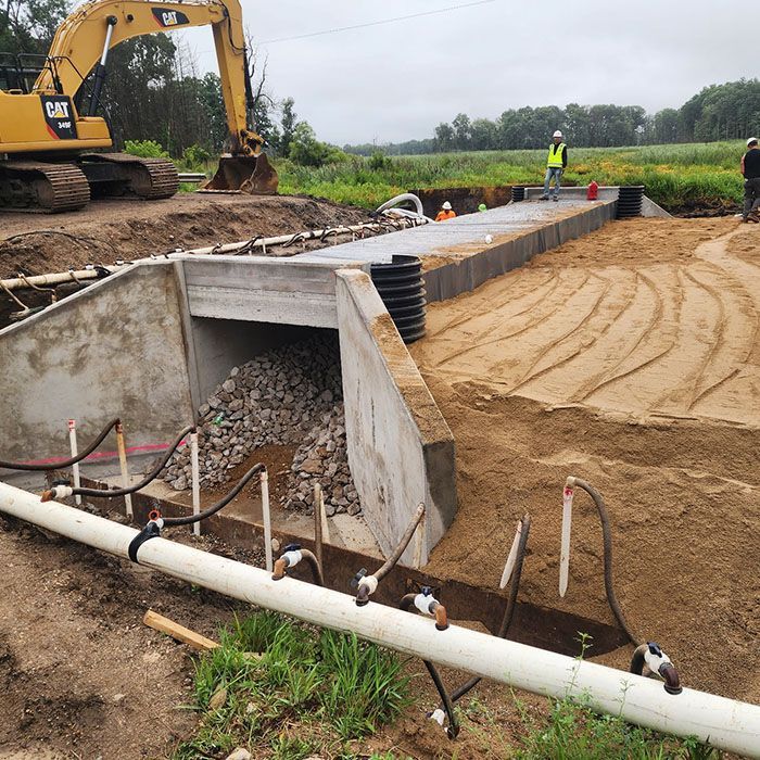 Construction site with excavator over concrete culvert and dirt embankment beside a pipe trench