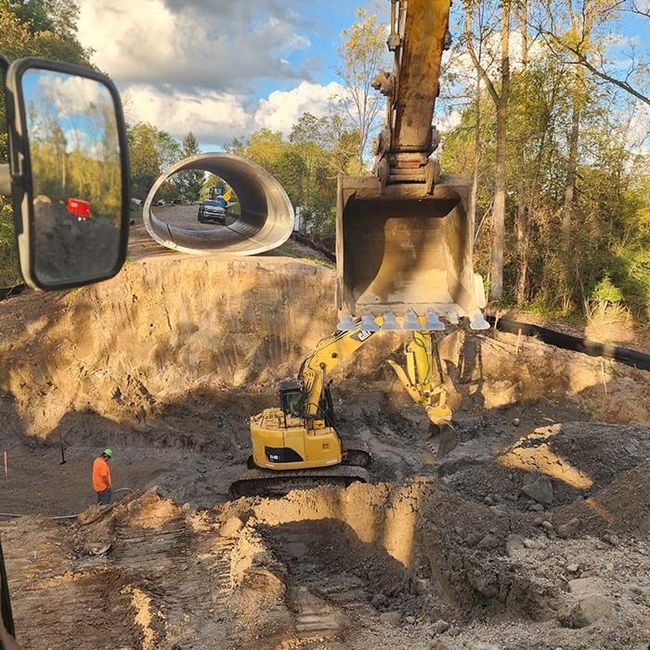 Yellow excavator digging a muddy construction pit, seen from inside a truck mirror; worker in orange nearby