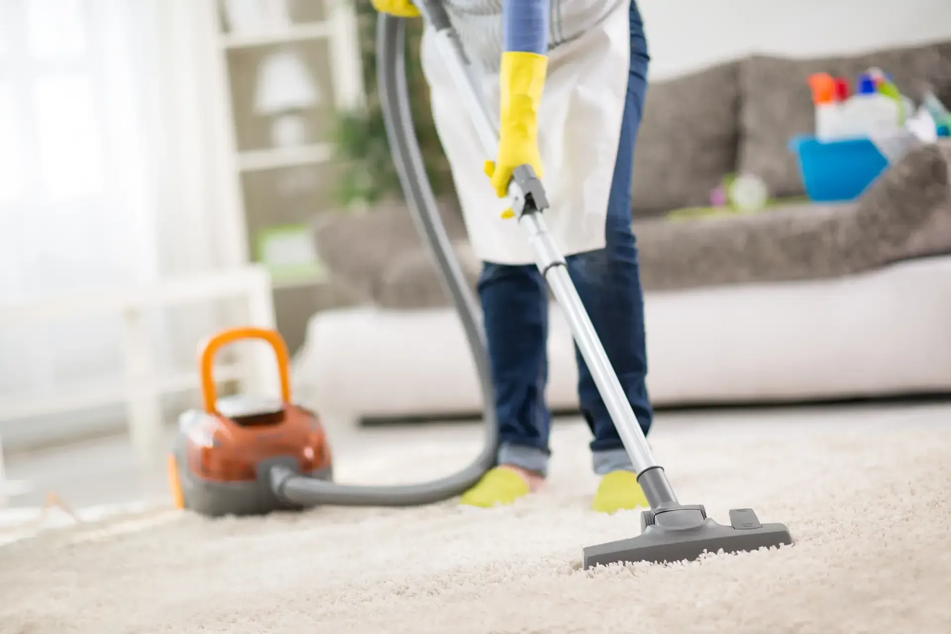Person vacuuming a carpet, wearing gloves and an apron, in a living room.