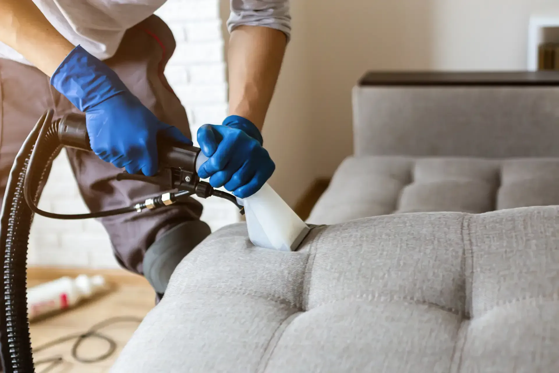 Person in blue gloves using upholstery cleaner on a gray tufted sofa.