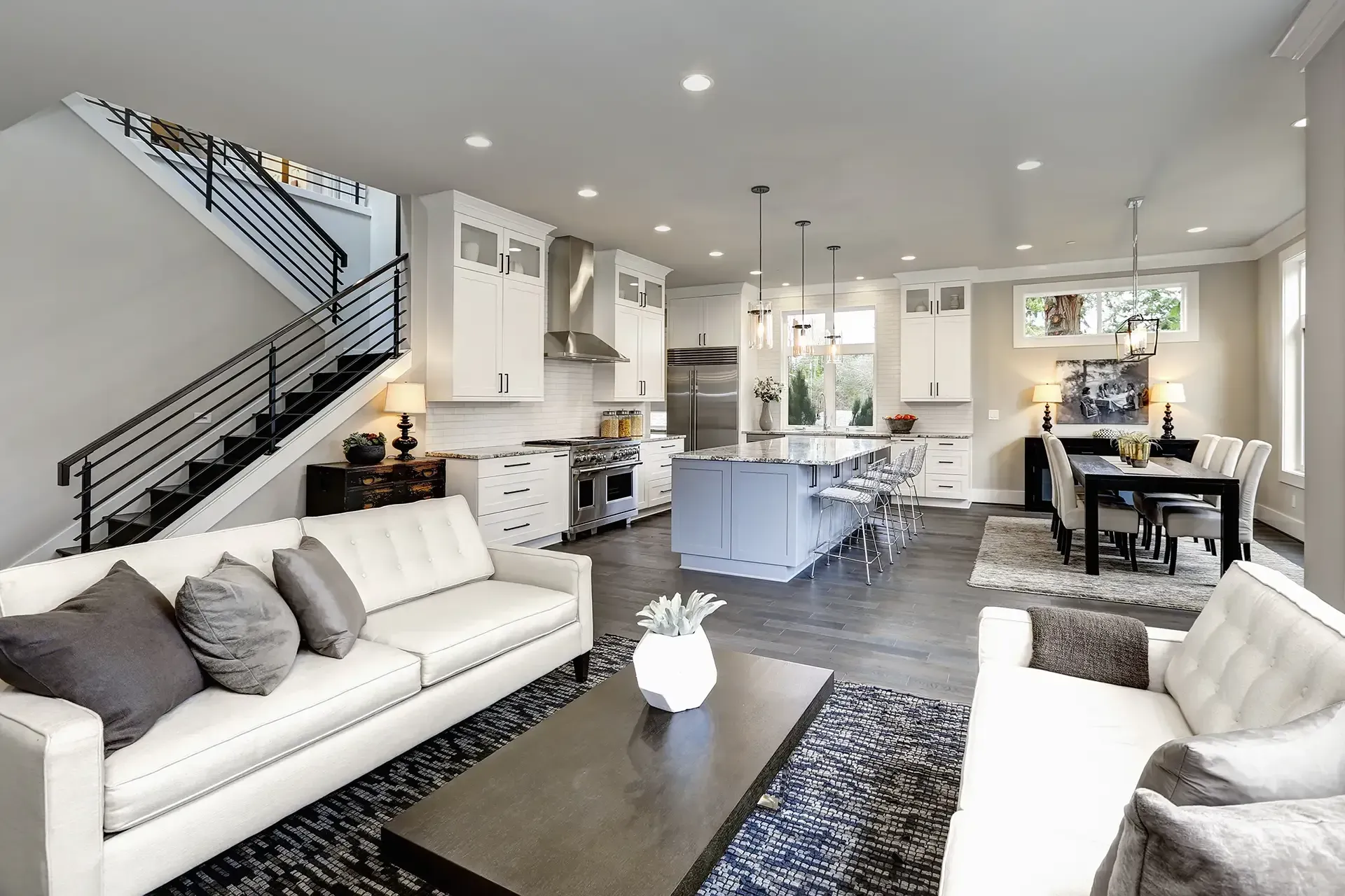 Modern open-concept living space with white kitchen, dining area, and sofas; a staircase is on the left.