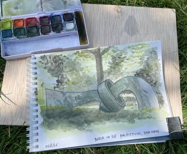 Aquarel schetsboektekening van een sculptuur onder een boom, met een aquarelpalet ernaast.