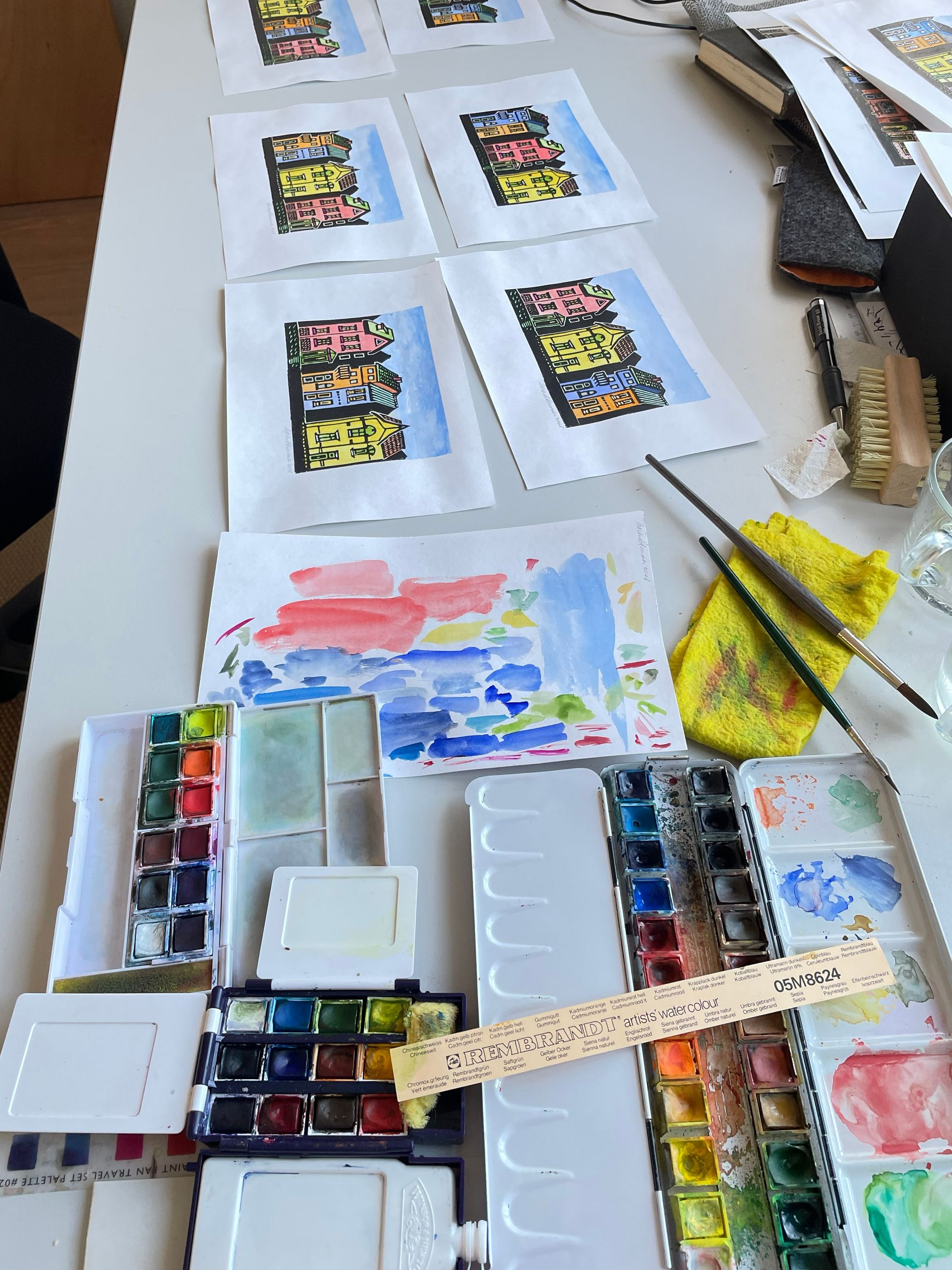 Op een wit bureau staan verschillende aquarellen van gebouwen, met verfpaletten, penselen en proefpapier in de buurt.