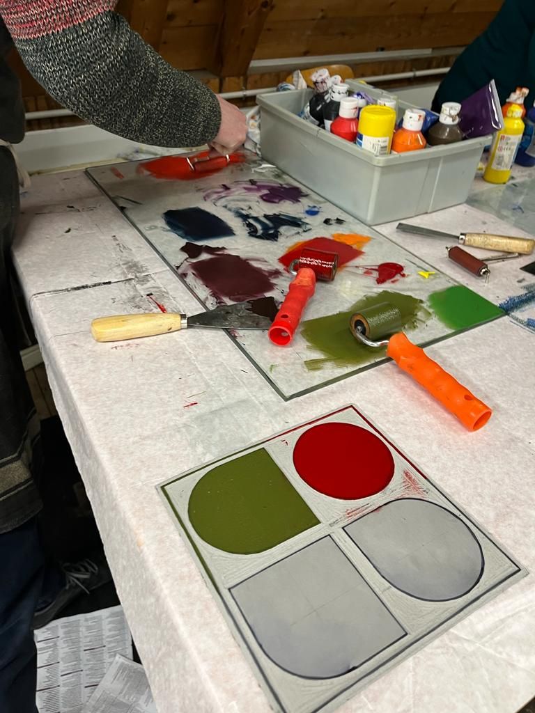 Een persoon gebruikt een drukrol op een glazen palet met kleurrijke inkt. Andere gereedschappen en verfpotjes zijn zichtbaar.