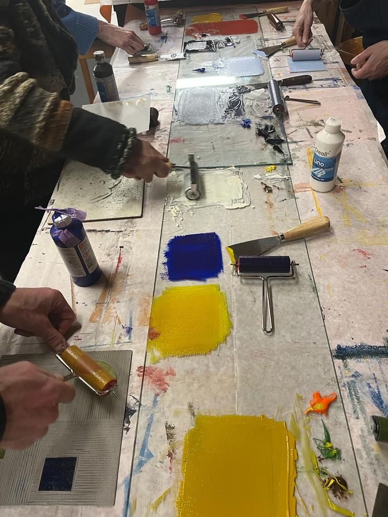 Mensen die aan een tafel druktechnieken toepassen met inktkleuren, rollers en gereedschap.