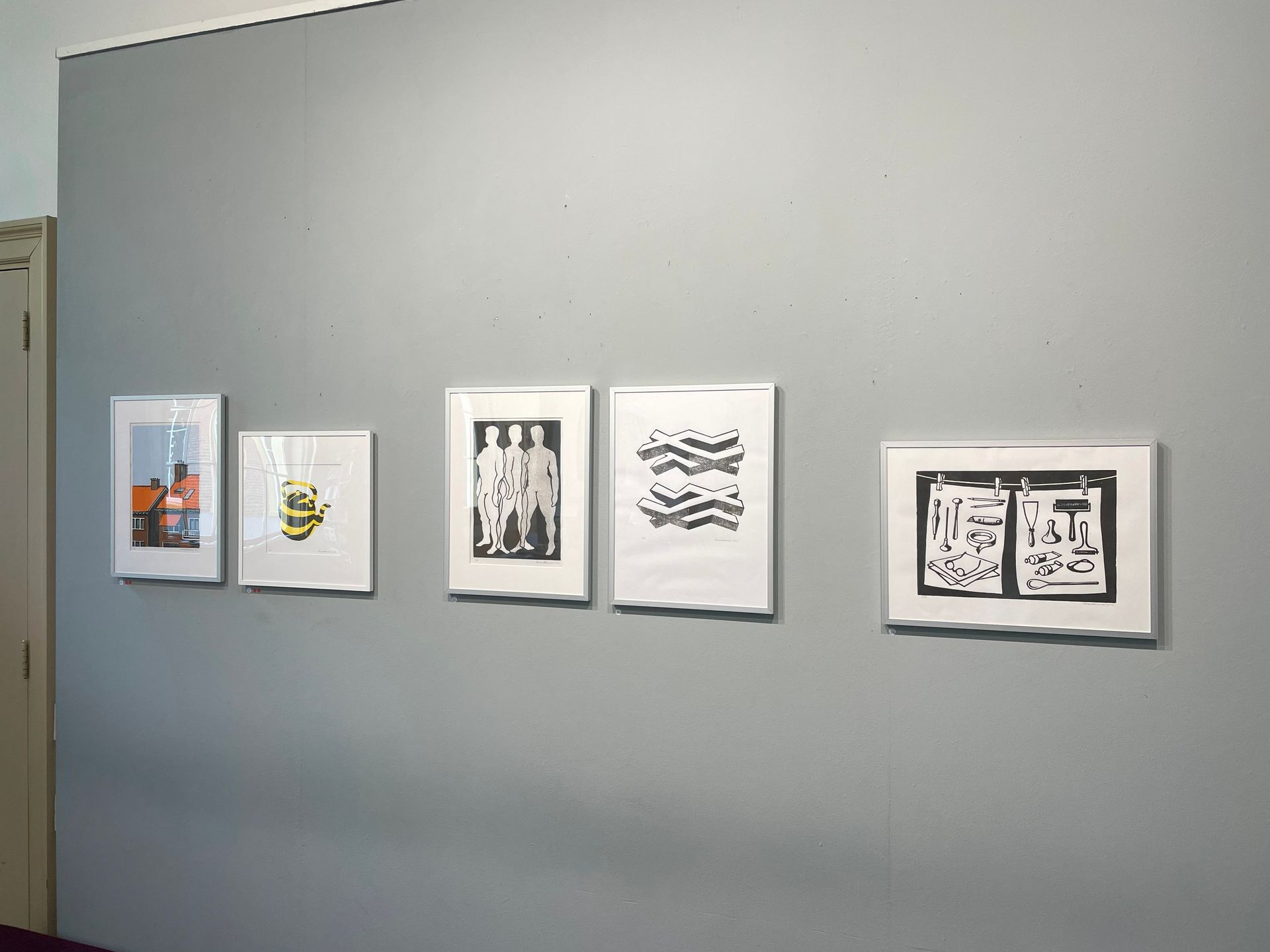 Aan een grijze muur hangen vijf ingelijste prenten. De prenten tonen verschillende zwart-witontwerpen.