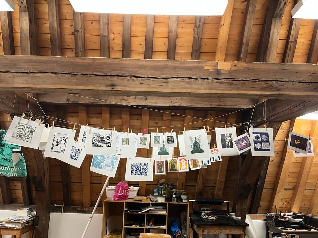 In het atelier hangen kunstprints aan een waslijn, met houten balken boven het hoofd.