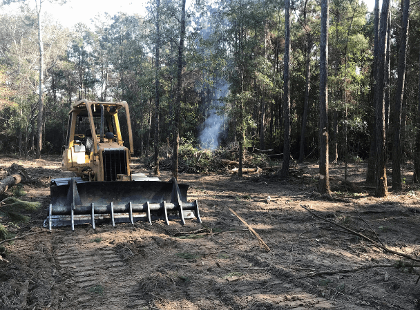 Bulldozer clearing trees in a forest, smoke visible in the background.