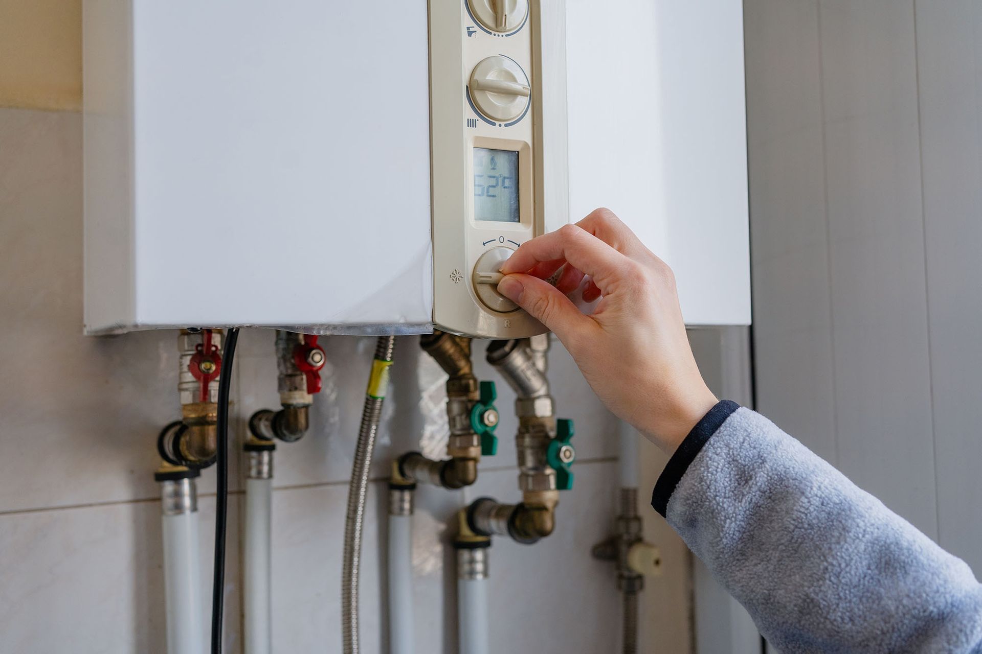 Hand turning a thermostat on a wall-mounted boiler with visible pipes and valves