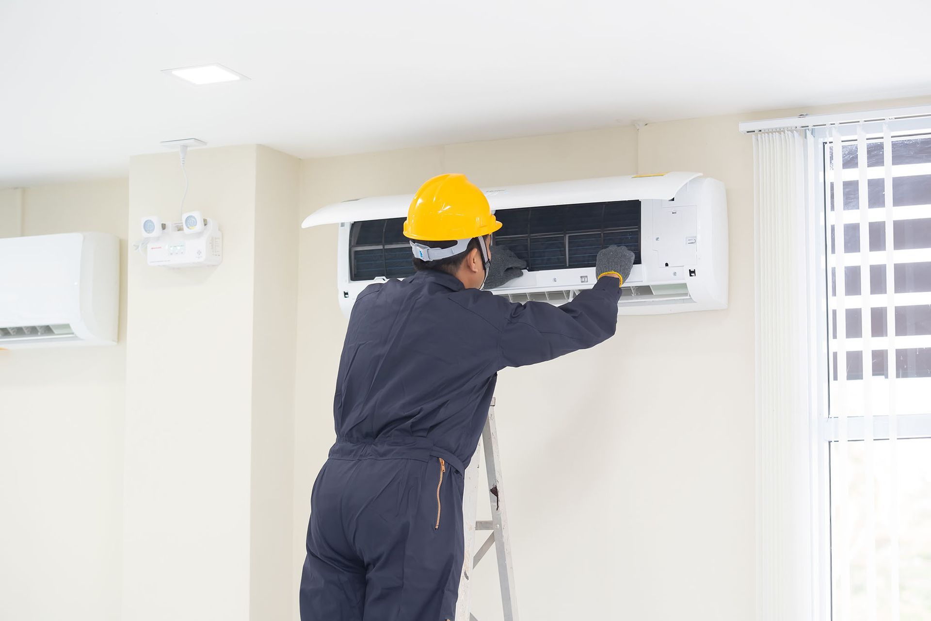 Technician in a yellow hard hat servicing a wall-mounted air conditioner indoors