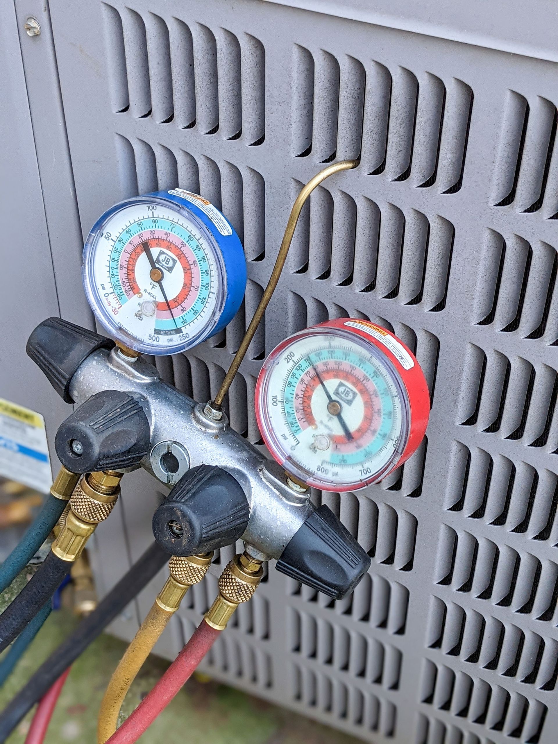 Two HVAC manifold gauges with red and blue dials connected to hoses on an outdoor unit.