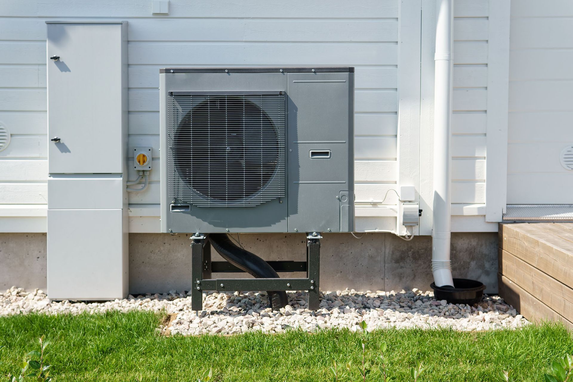 Outdoor HVAC unit beside a white house, with a utility box and downspout on a gravel bed near grass