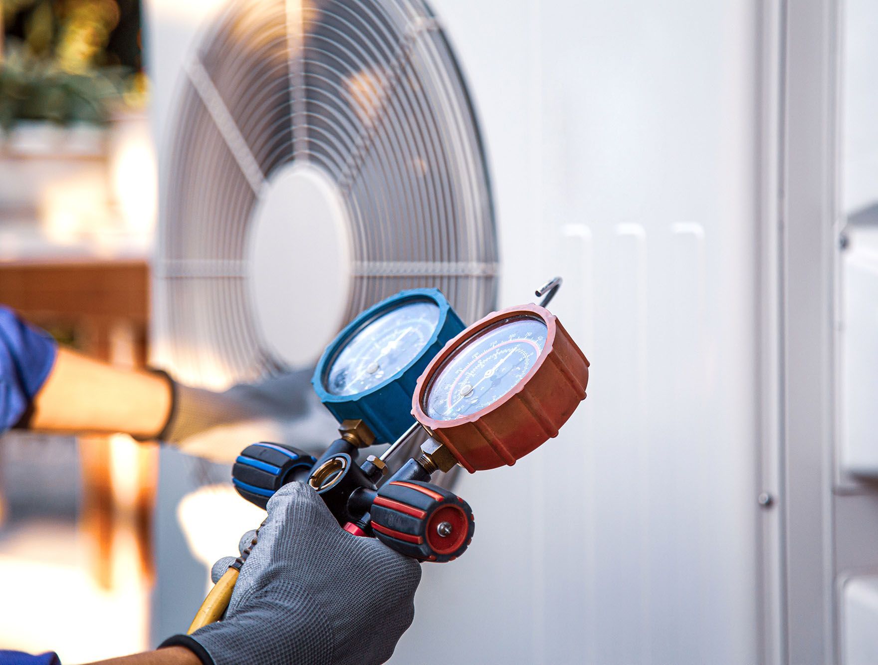 Gloved hand holding HVAC gauges beside an outdoor air conditioner unit and spinning fan