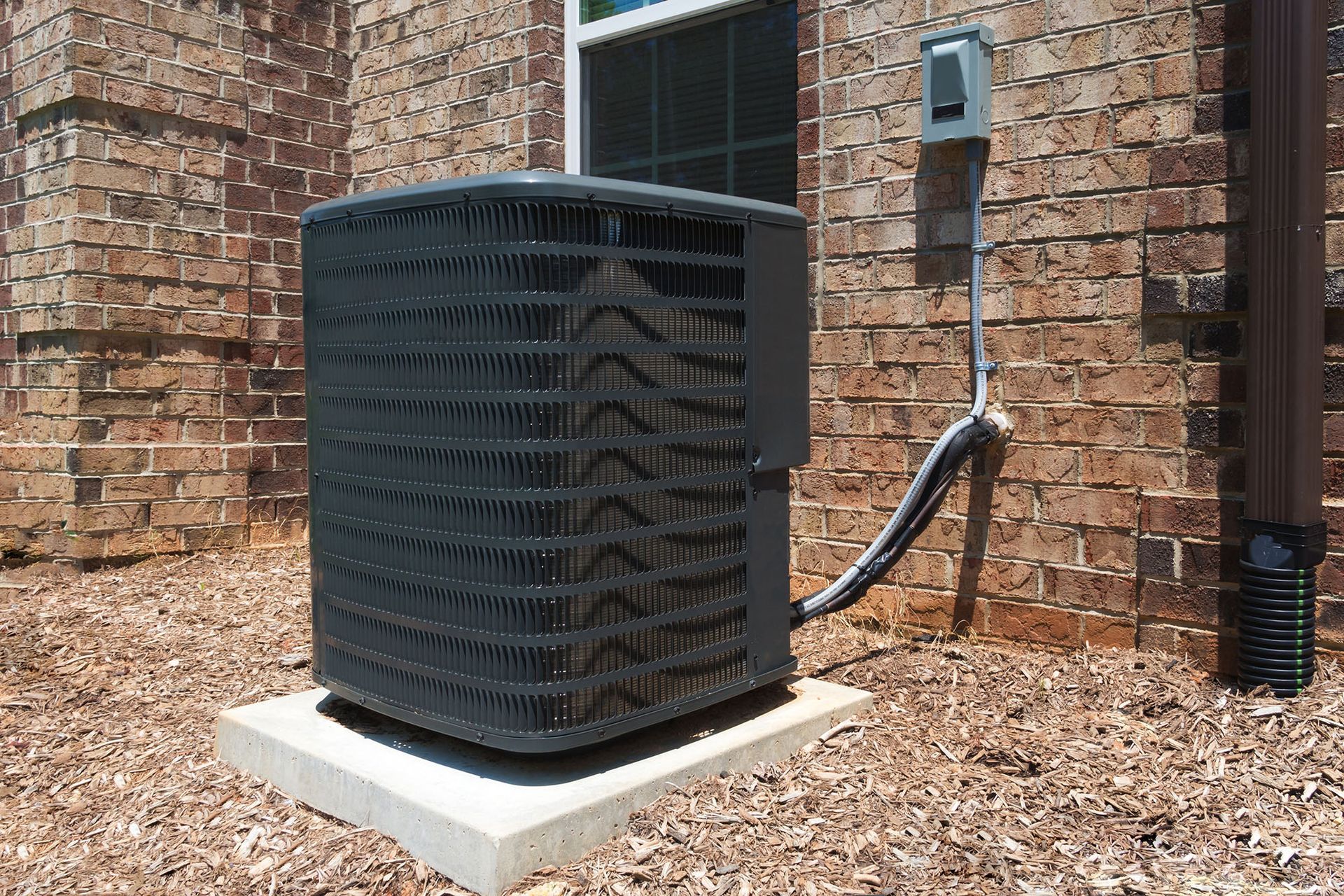 Outdoor air-conditioning unit beside a brick house, with a black hose and concrete pad.