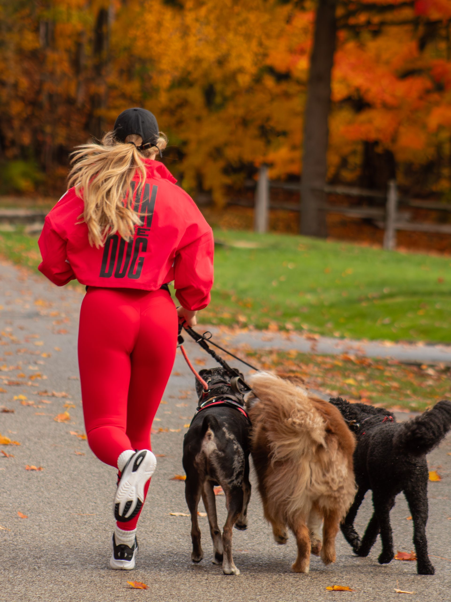 Woman in red running with three dogs on a path, fall foliage in the background.