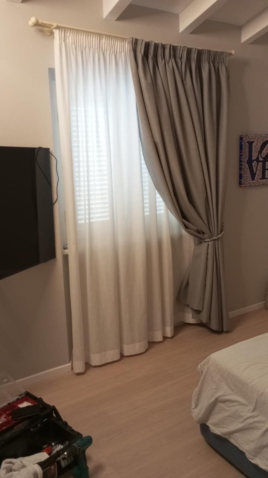 Finestra della camera da letto con tende bianche trasparenti e drappeggi grigi, accanto a un letto sulla destra.