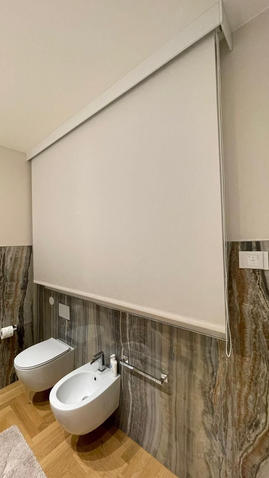 Bagno accessibile con WC a parete, bidet e grande tenda bianca sopra la finestra