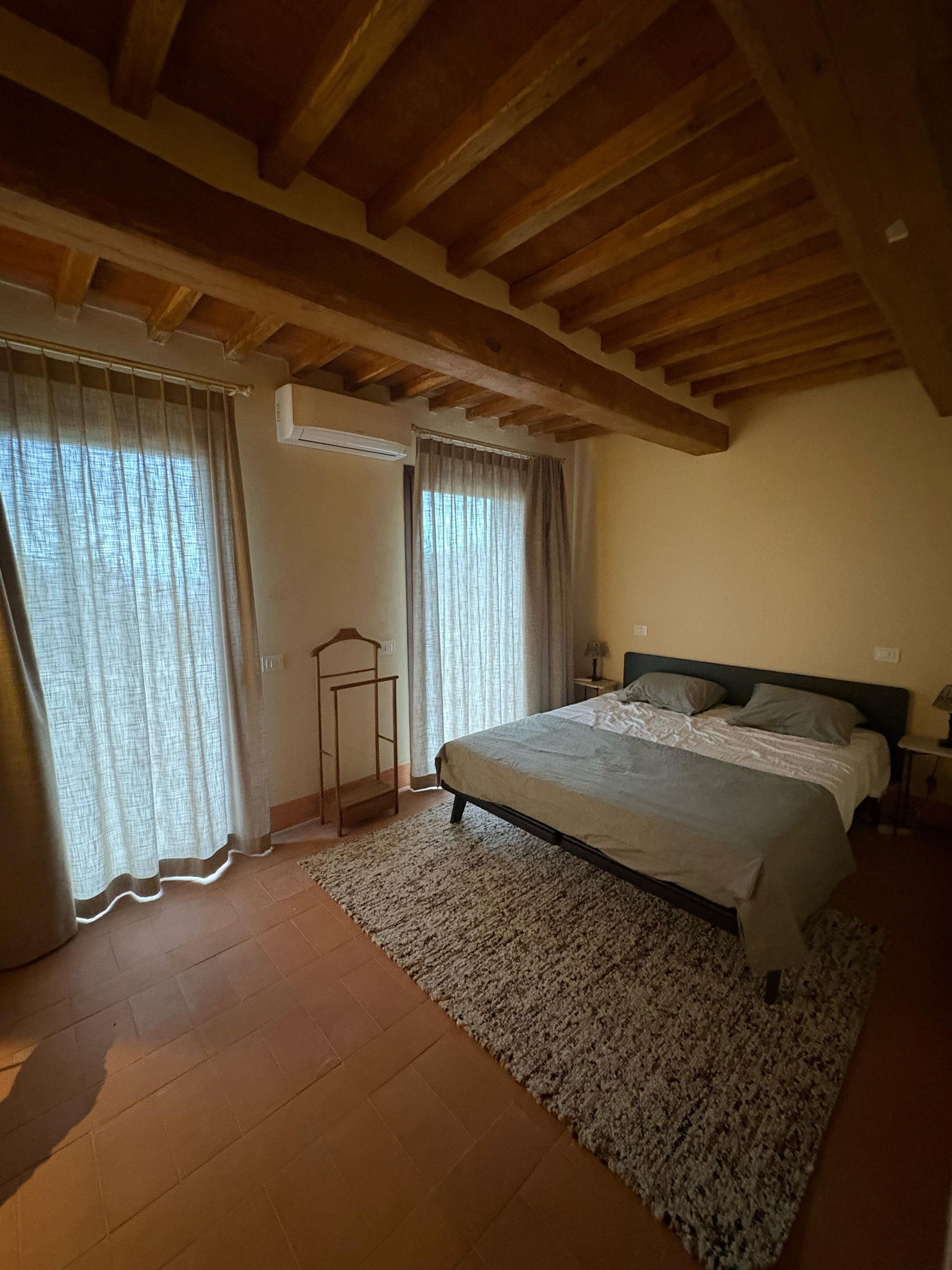 Accogliente camera da letto con soffitto con travi a vista, letto, tappeto e tende trasparenti che lasciano entrare una luce soffusa.