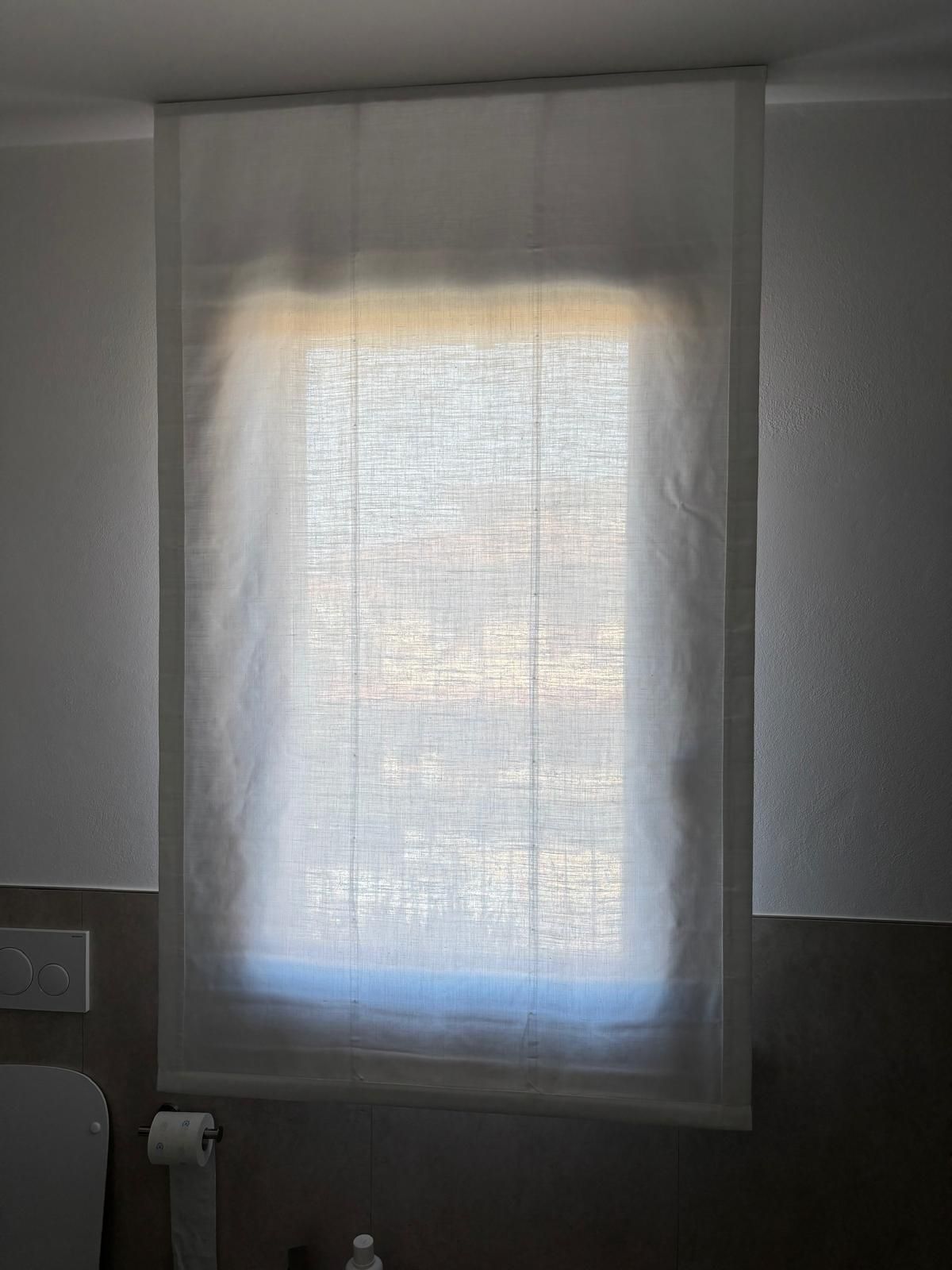Una tenda bianca trasparente copre una finestra da cui filtra la luce del giorno in una stanza grigia.
