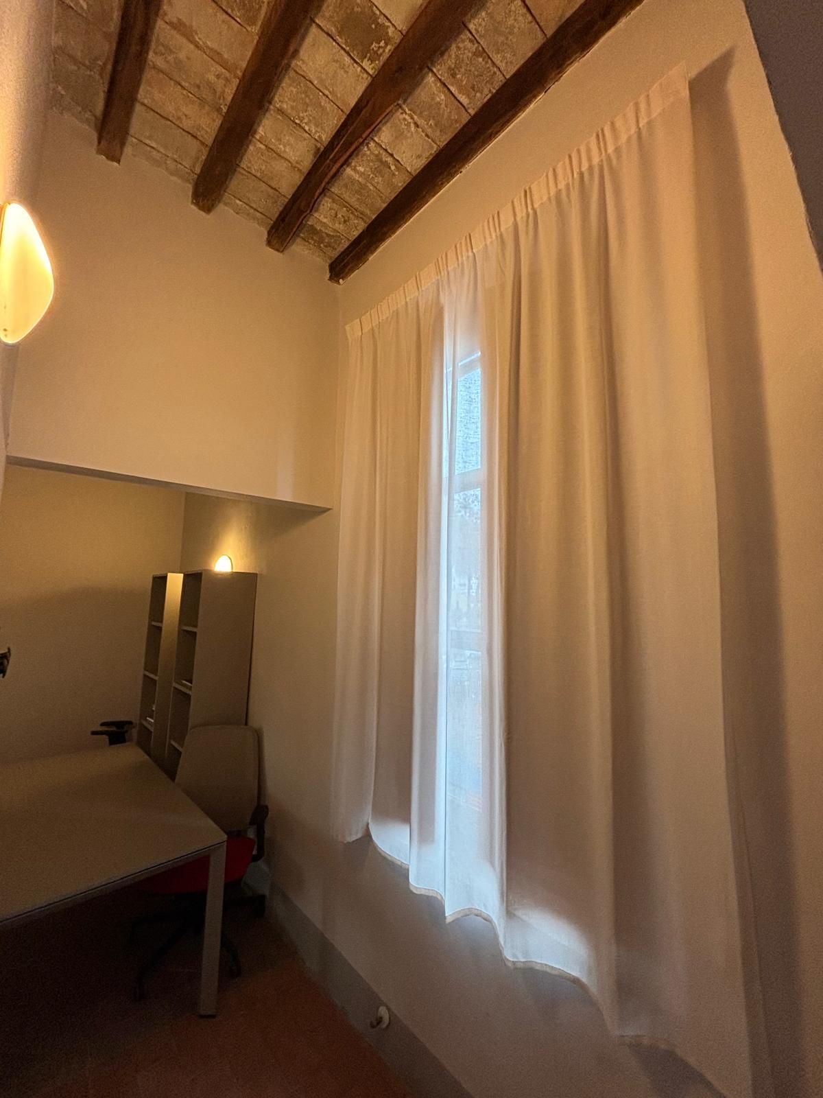 Camera da letto con soffitto alto, travi a vista, letto e ampia finestra con tende che lascia entrare una luce soffusa.