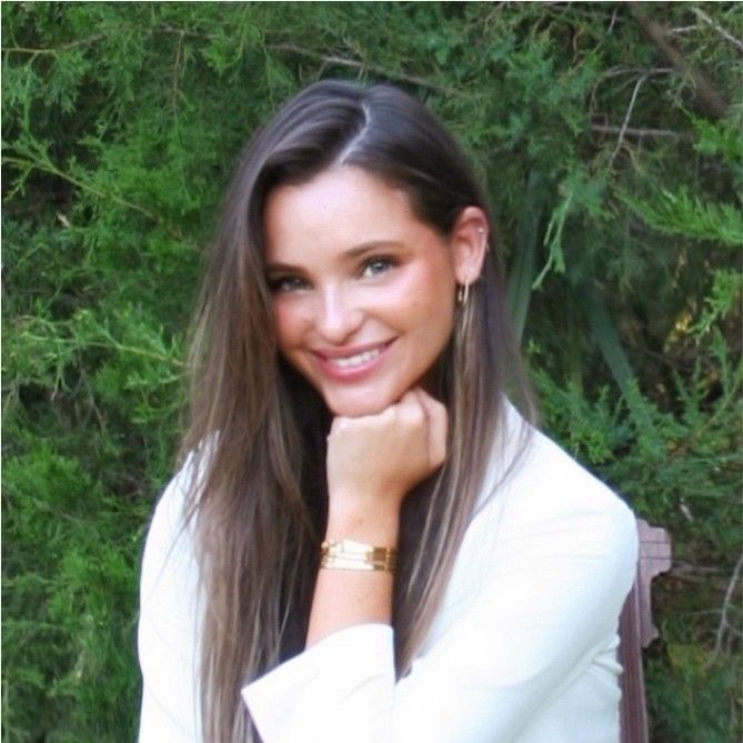 A person smiling with a hand resting on their chin, wearing a white blazer against a backdrop of green foliage.