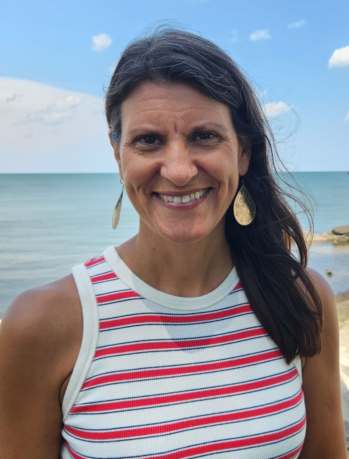 A smiling person wearing a red and white striped tank top and gold teardrop earrings stands before a calm blue sea.