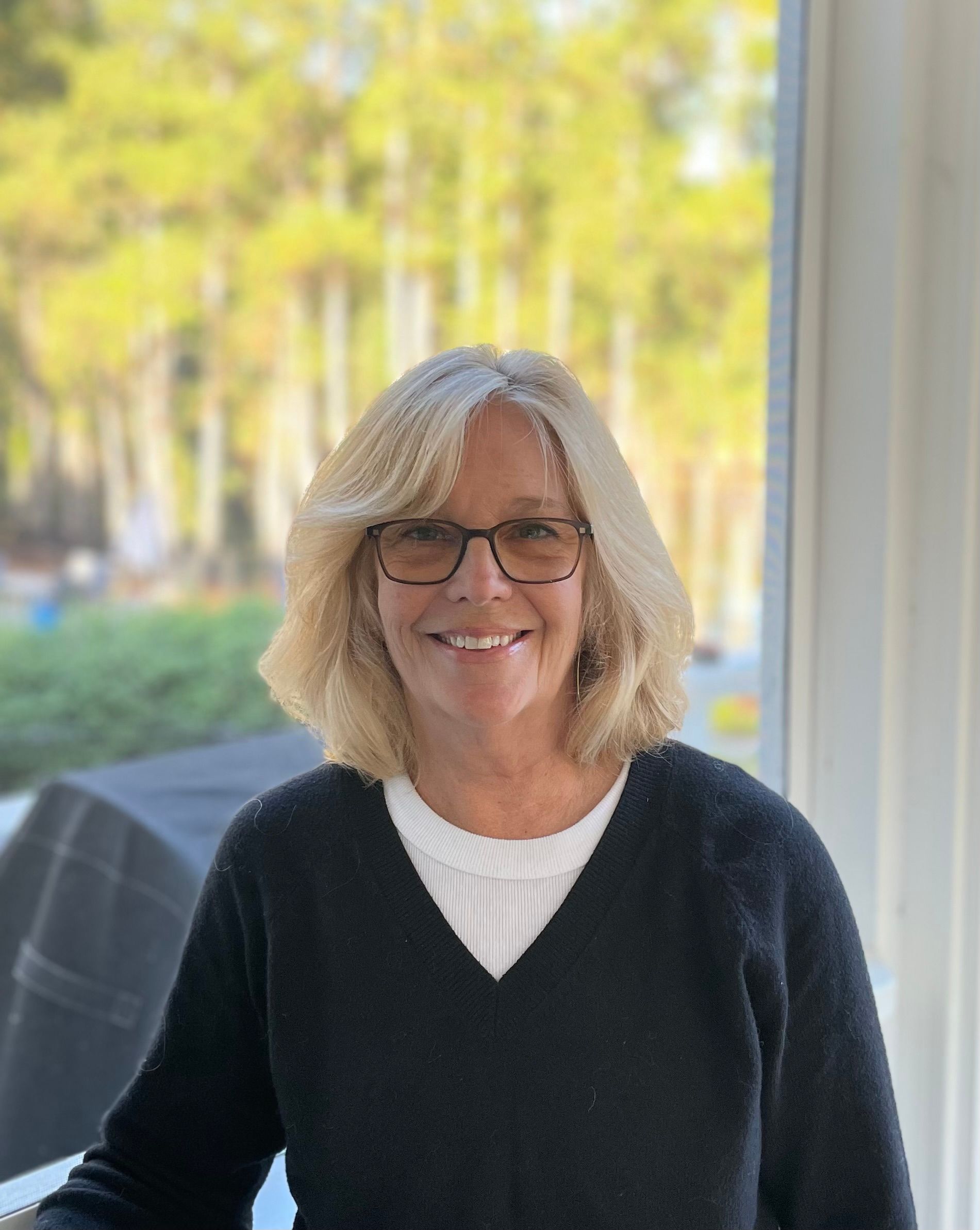 A person with shoulder-length blonde hair and glasses wearing a black sweater and white shirt, smiling indoors.