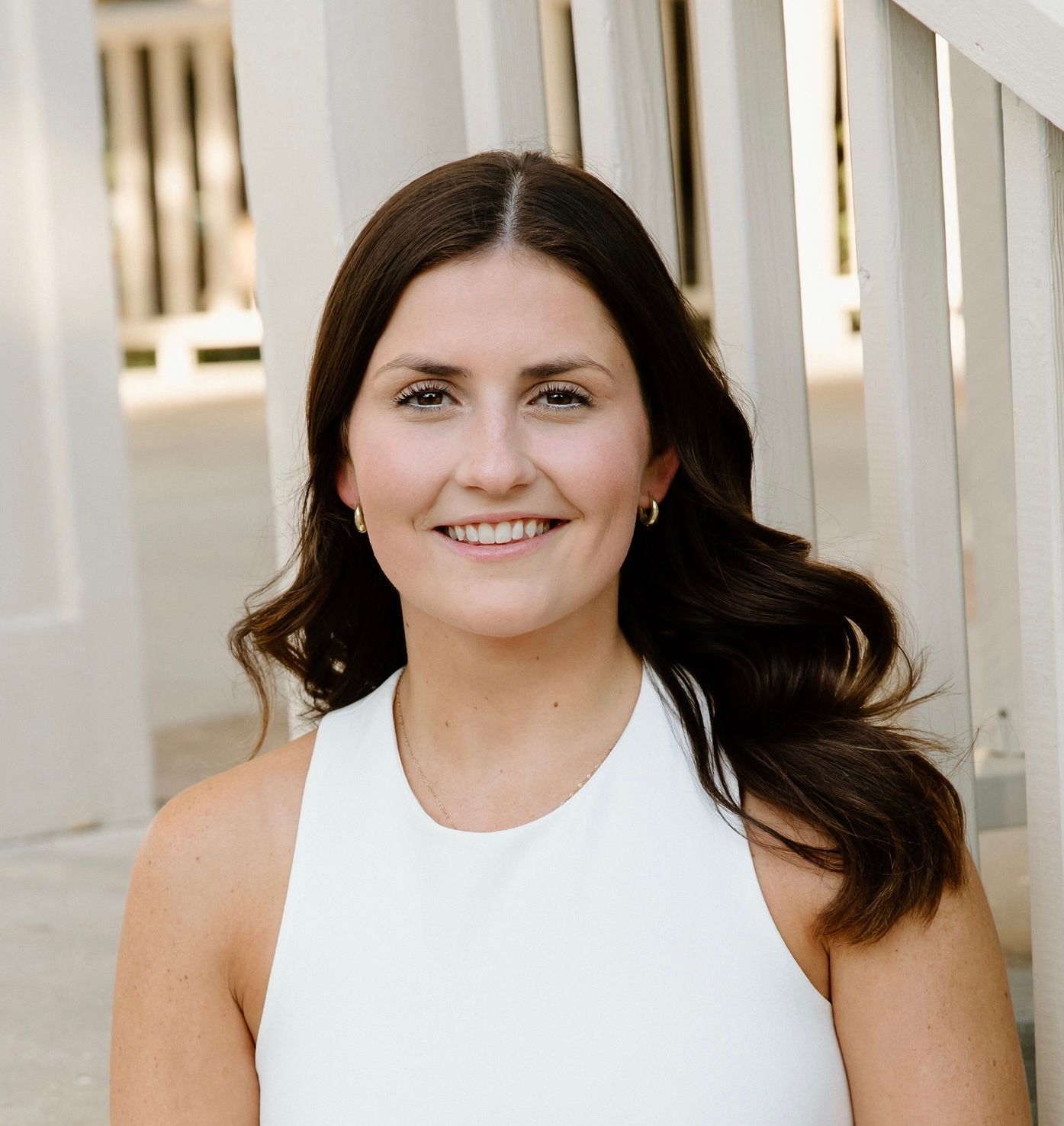 A smiling woman with dark hair wearing a sleeveless white top, posing outdoors in front of a white wooden structure.