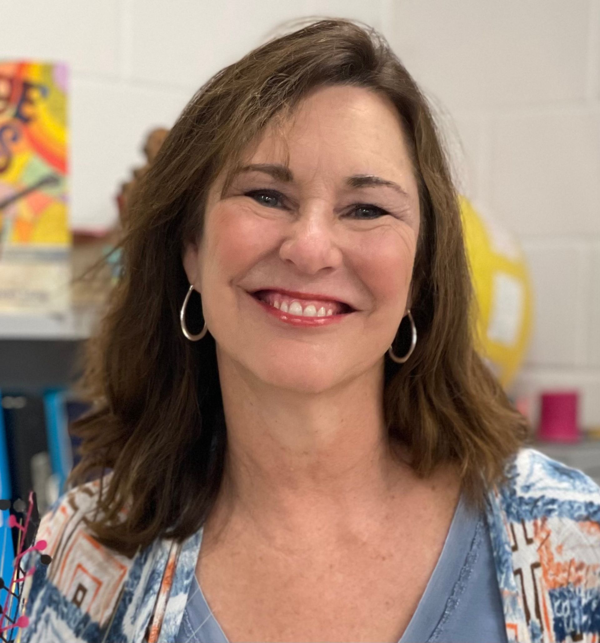 A smiling person with shoulder-length brown hair and hoop earrings, posing in front of a shelf with books and supplies.