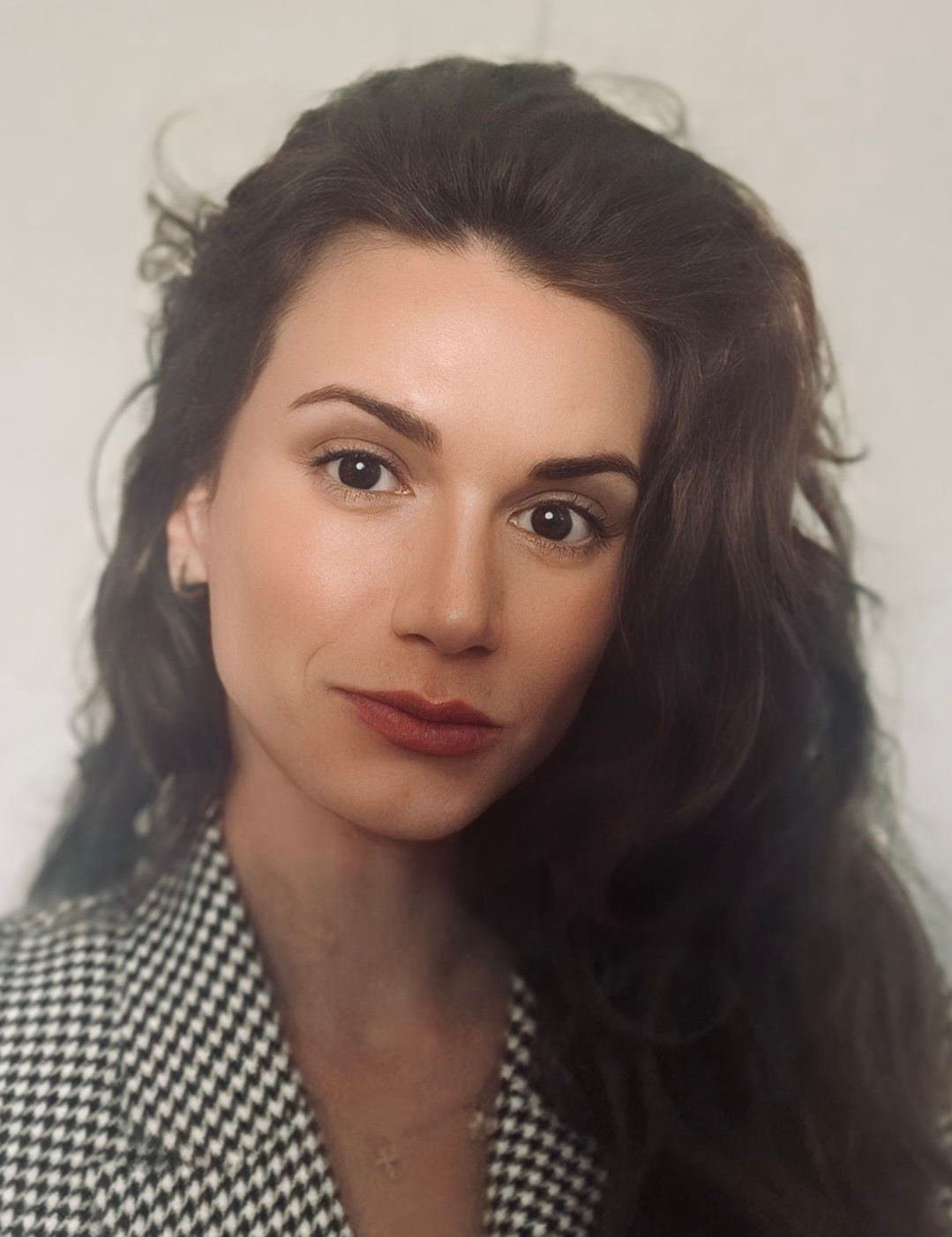 A headshot of a person with long dark hair, wearing a black and white houndstooth blazer, looking toward the camera.