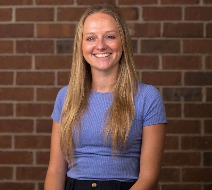 A person with long blonde hair smiling, wearing a blue shirt, standing in front of a brown brick wall.